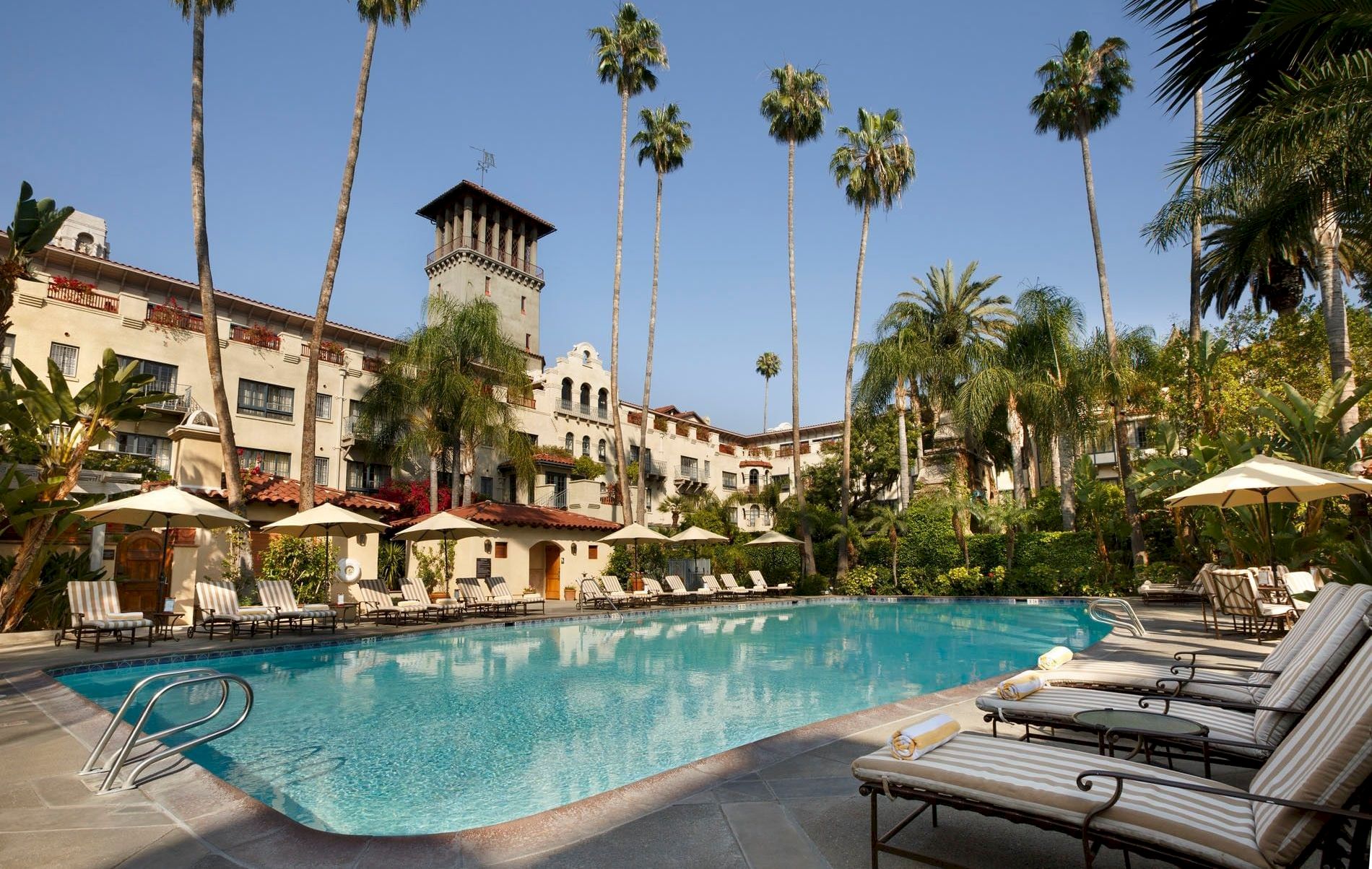 A sunny hotel courtyard with a swimming pool, striped loungers, umbrellas, and tall palm trees, with a castle-like tower building.