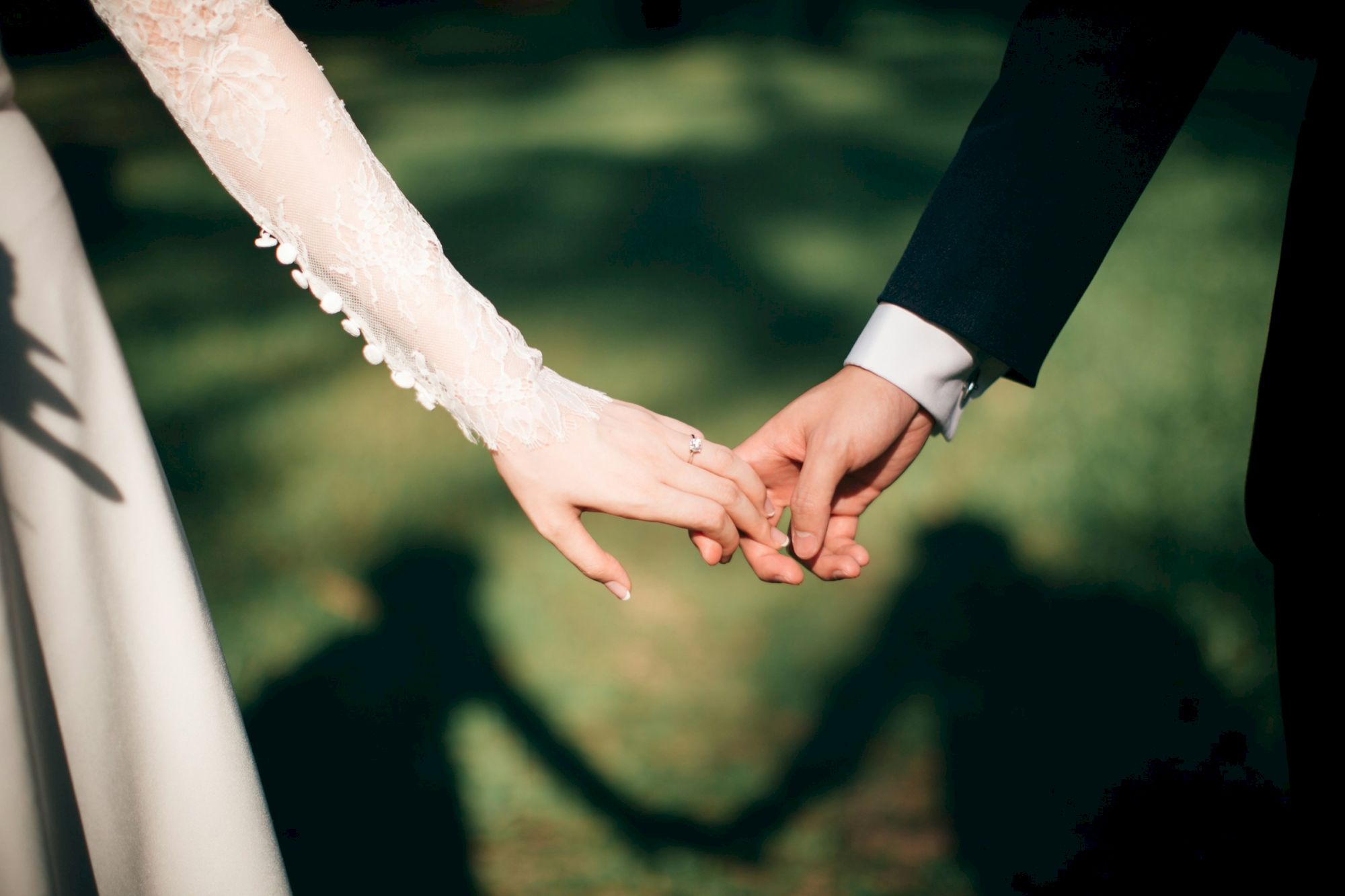 A bride and groom hold hands outdoors, with her lace veil sleeve and his suit cuff, sharing a tender wedding moment.