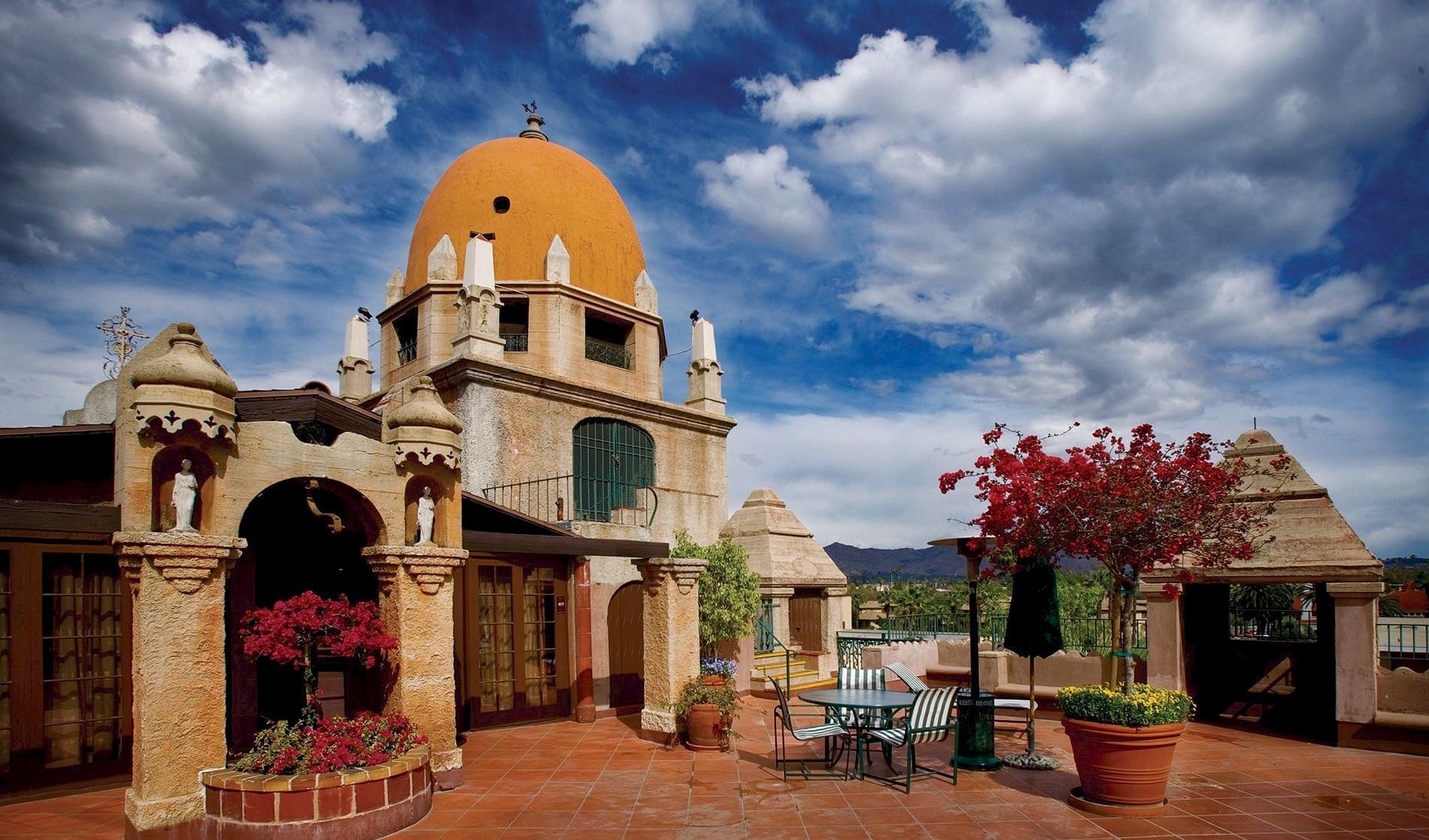 A yellow-domed church in a sunny courtyard, with red flowers and patio seating beneath dramatic blue clouds.