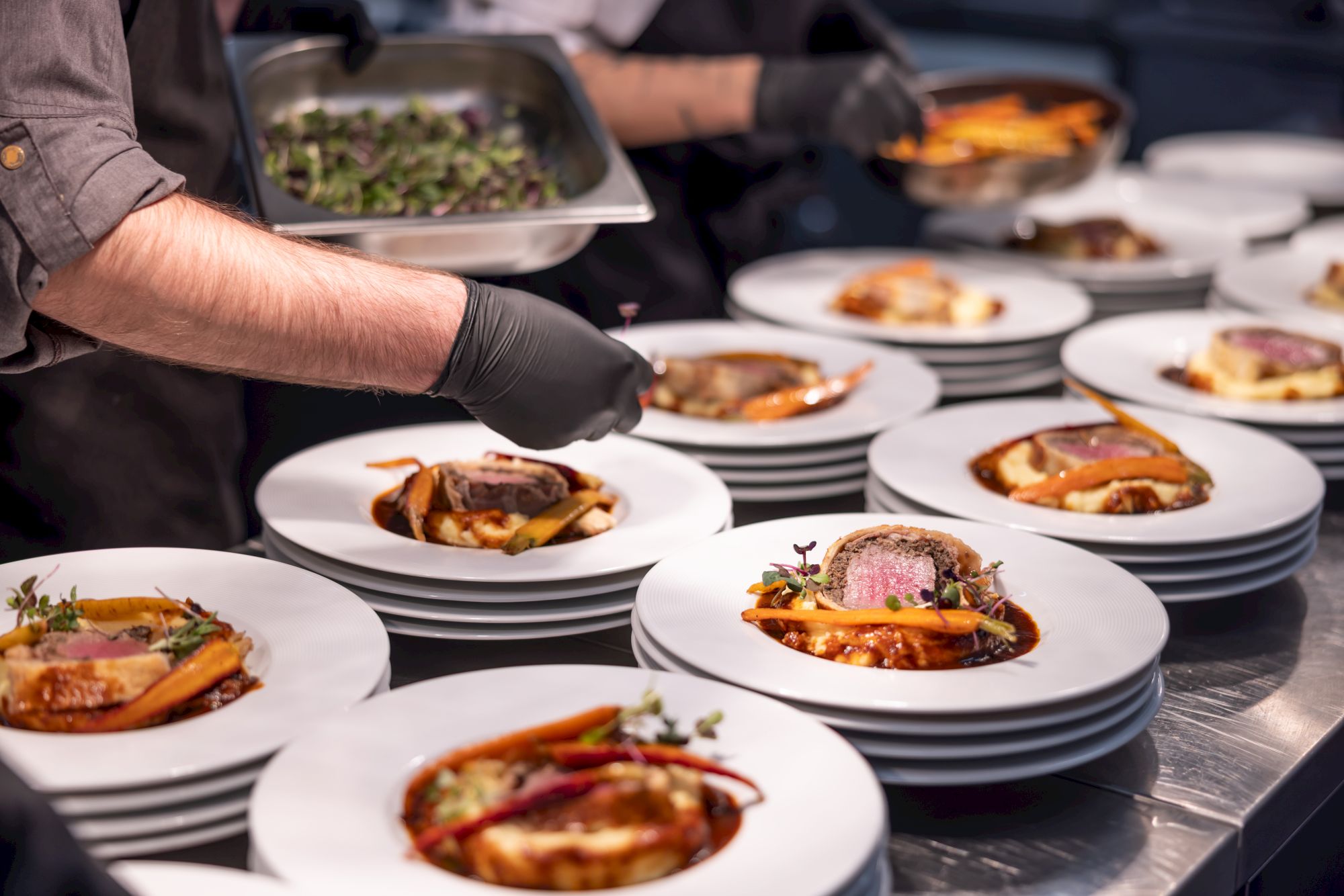 A chef in gloves plates gourmet dishes on white plates during a catering service, with trays of food in the background.