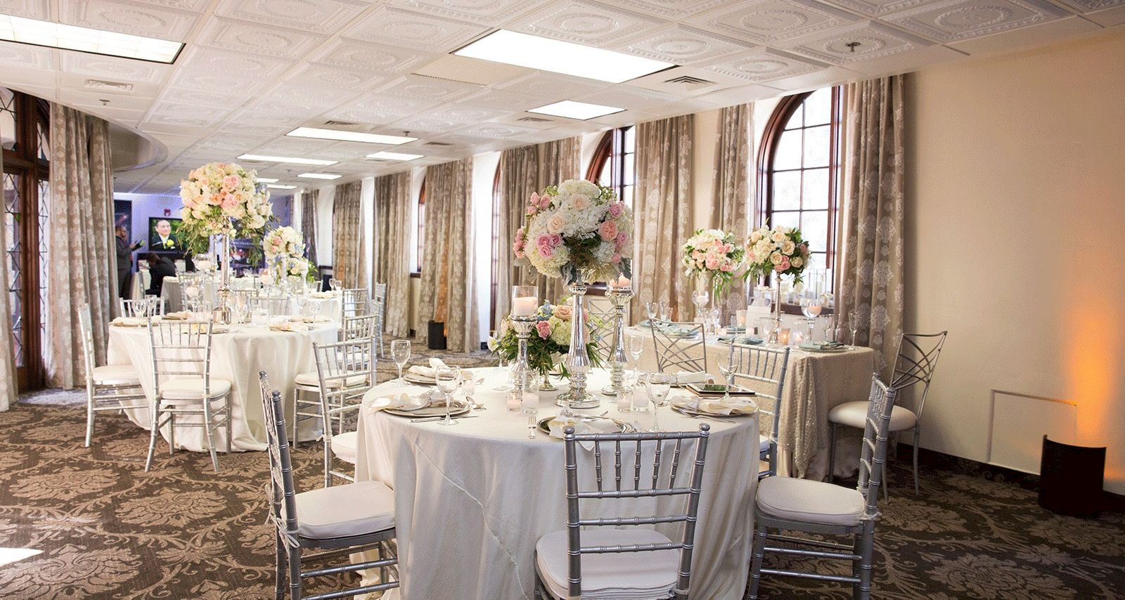An elegant event hall set for a wedding or banquet, with round tables, silver chairs, and tall floral centerpieces by tall windows.