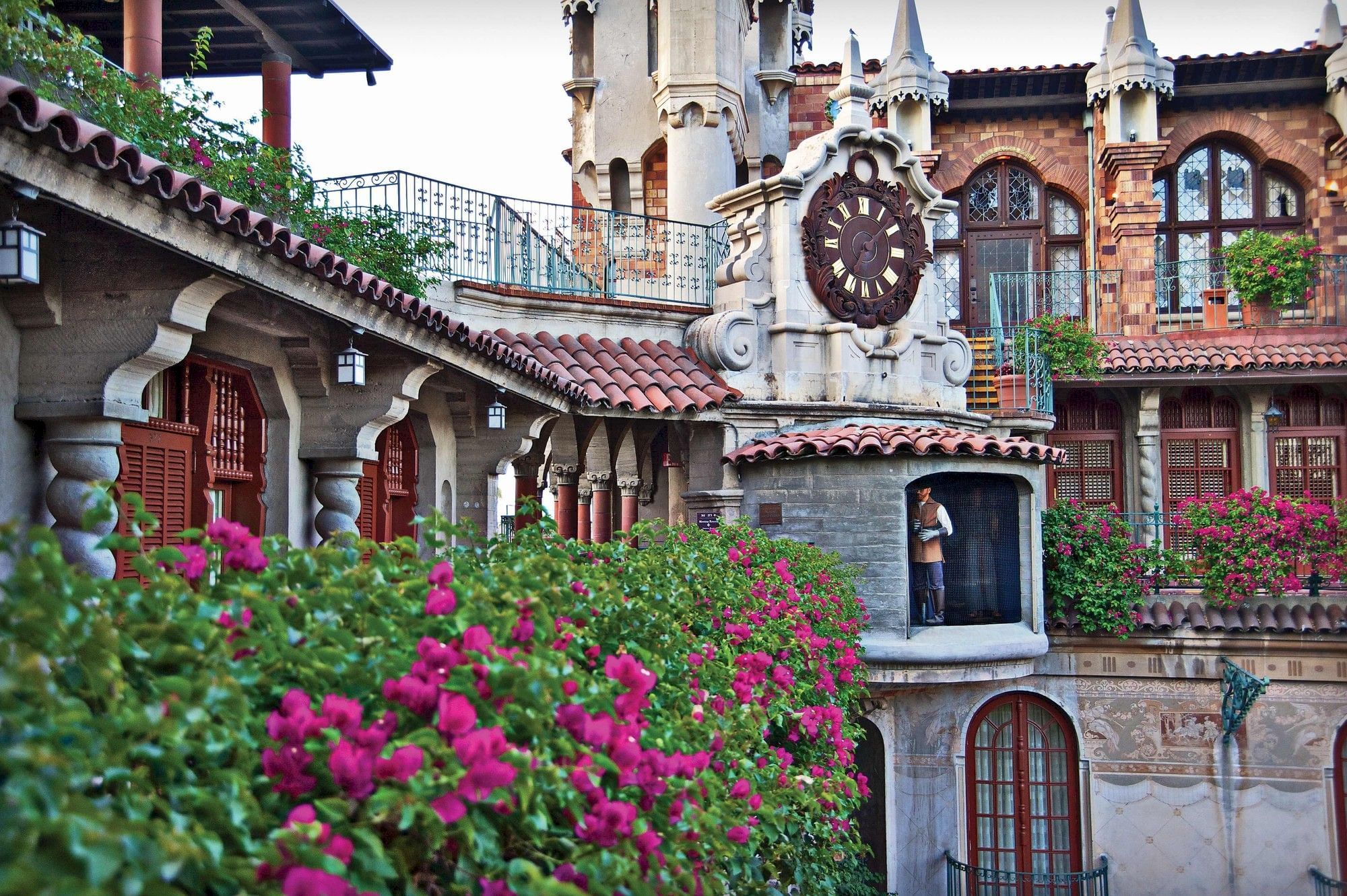 An ornate stucco clock tower with Roman numerals stands beside arched windows, framed by lush pink flowers in the foreground.