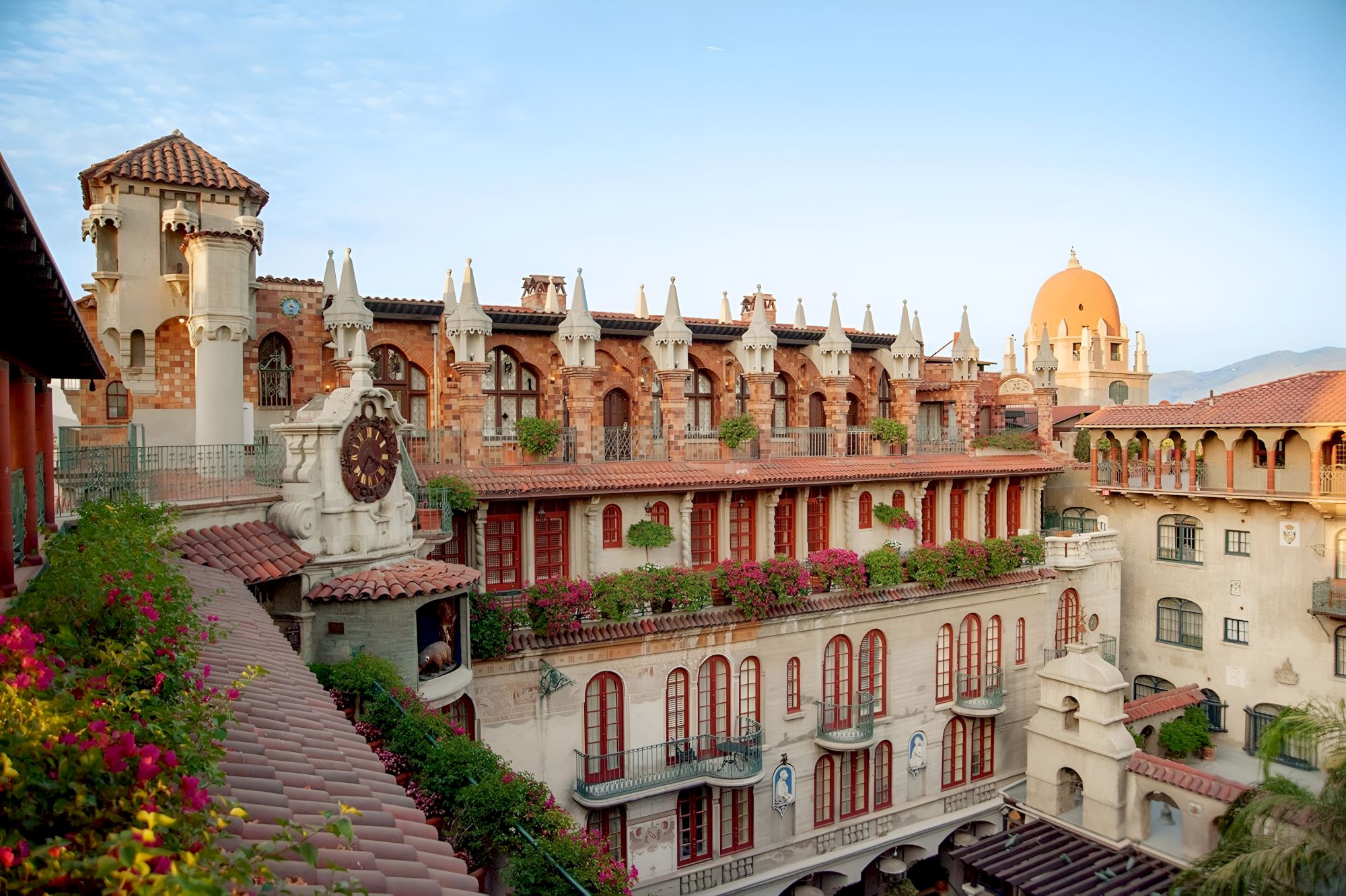 Spanish-style complex with arched corridors, white towers and domes, and colorful flowers under a blue sky.