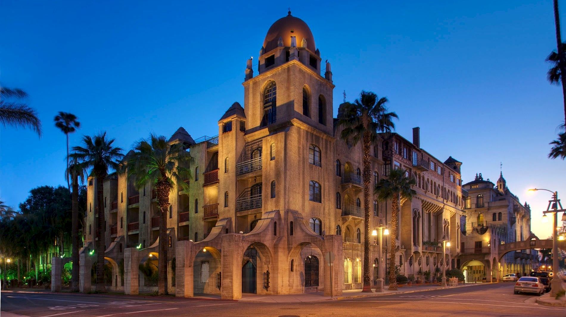 A grand hotel with a lit domed tower rises above palm trees at dusk, with streetlights and cars along the road.