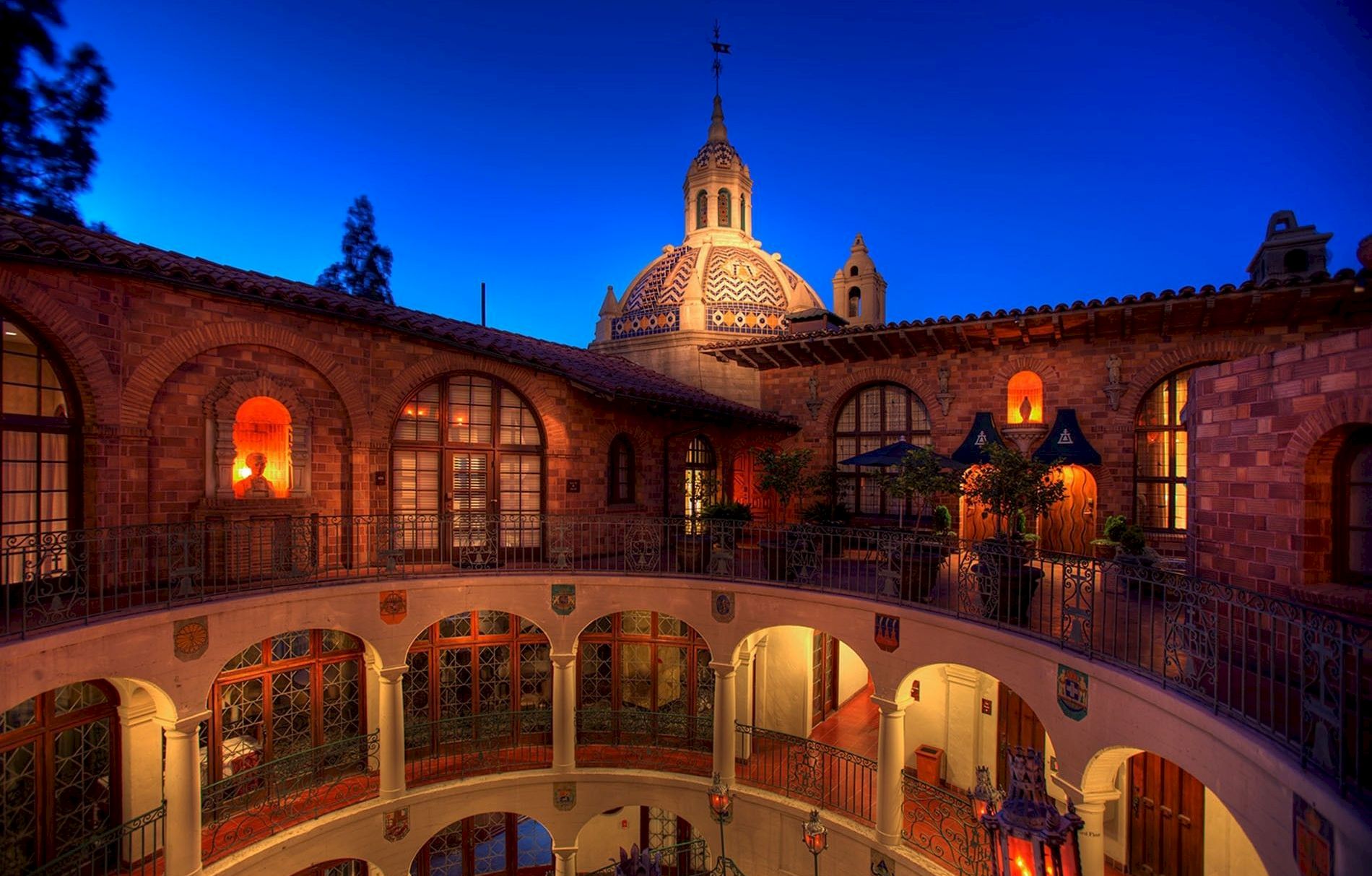 A night view of a brick courtyard with arches and glowing lamps, featuring a central domed tower under a deep blue sky.