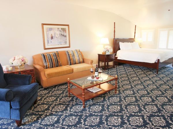 A bright hotel living room with a sofa, coffee table, and a neatly made bed by sunny windows.