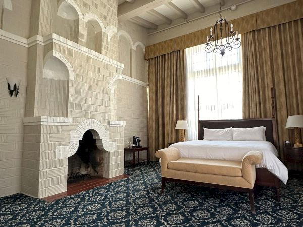 An elegant hotel bedroom with a brick fireplace, chandelier, large bed, patterned rug, and tall curtains under arched beams.