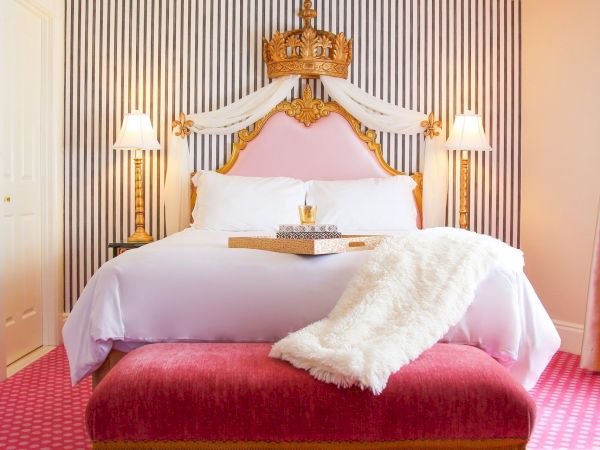 Luxurious hotel bedroom with a gold crown canopy bed, white linens, lamps, striped wall, and a plush throw on pink bedding.