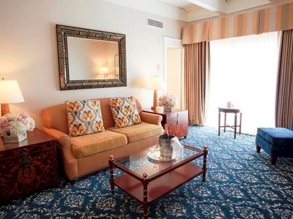 A bright hotel living room with a sofa, lamps, coffee table, floral d&eacute;cor, and blue carpet leading to a sunlit window.