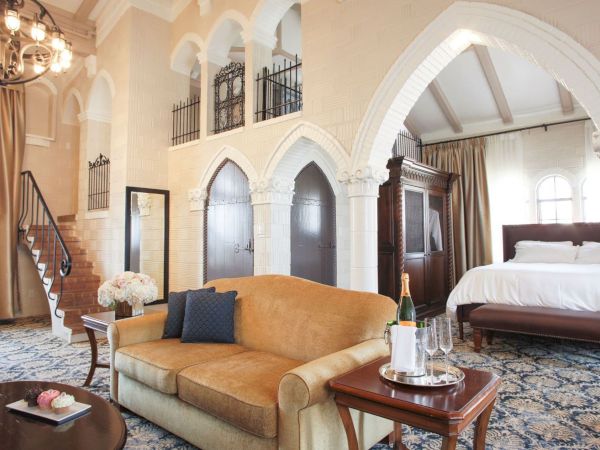 An elegant hotel suite with an arched white interior, warm chandeliers, a gold sofa, a bed, and a sparkling table set.