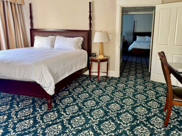 Hotel room with a dark wooden bed, white pillows, bedside table with lamp, and blue patterned carpet by a doorway to another room.