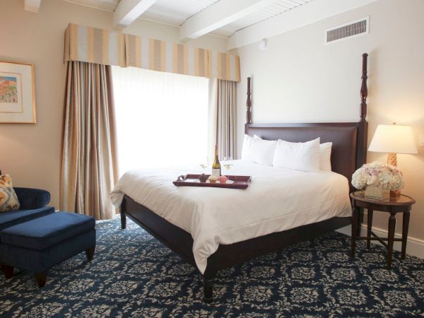 A cozy hotel bedroom with a king bed, curtains, bedside lamps, a side table with flowers, and a lounge chair.