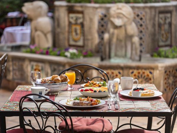 An outdoor patio table set for breakfast with coffee, juice, pastries, and fruit in front of an ornate bench backdrop.