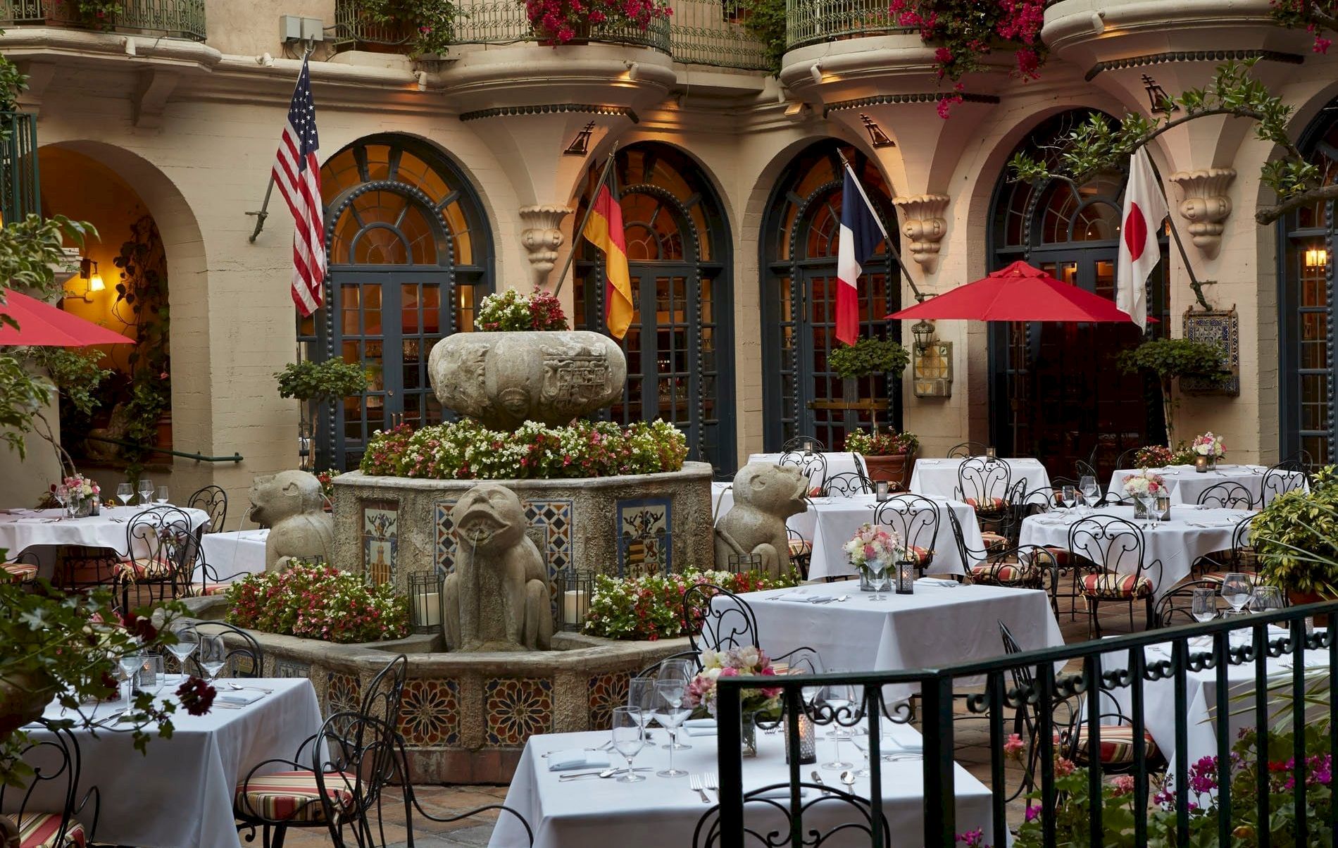An outdoor courtyard dining setup with white tablecloths, flowers, statues, and flags of the USA, Germany, and Japan.