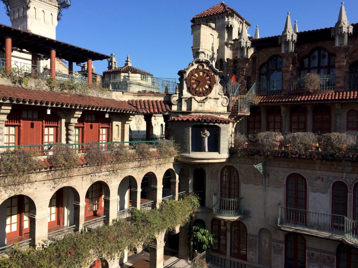 A sunny courtyard with ornate historic Spanish-style buildings, arched walkways, red shutters, and a decorative clock fountain in view.
