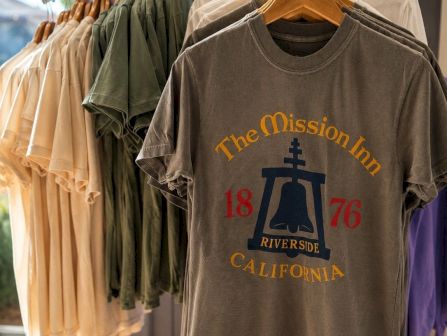Clothing rack with a graphic T-shirt: &ldquo;The Mission Inn,&rdquo; &ldquo;Riverside, California,&rdquo; year &ldquo;1876,&rdquo; on wooden hangers.