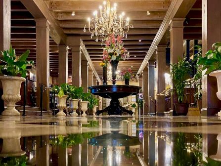 An elegant indoor hall with a crystal chandelier, floral centerpiece, and a reflective fountain basin between tall columns.