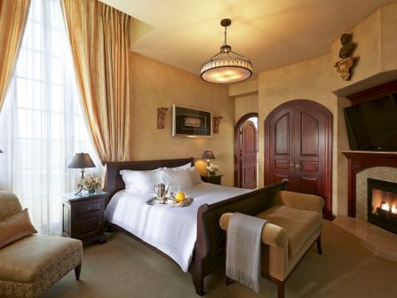 A luxurious hotel bedroom with a bed set, chandelier, ornate doors, tall curtains, and a glowing fireplace.