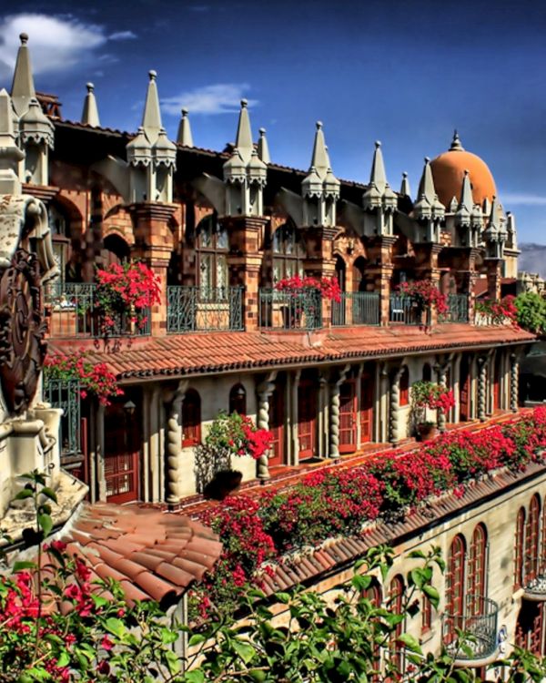 Ornate historic courtyard with brick arches, white spires, and lots of red flowers under a bright blue sky.