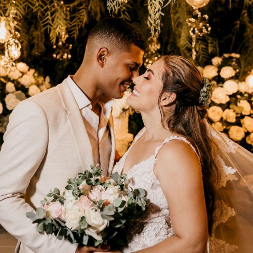 A bride and groom share a close kiss at a warm, fairy-lit wedding backdrop, surrounded by flowers and greenery.