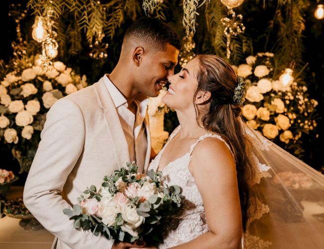 A bride and groom share a close kiss at a warm, fairy-lit wedding backdrop, surrounded by flowers and greenery.