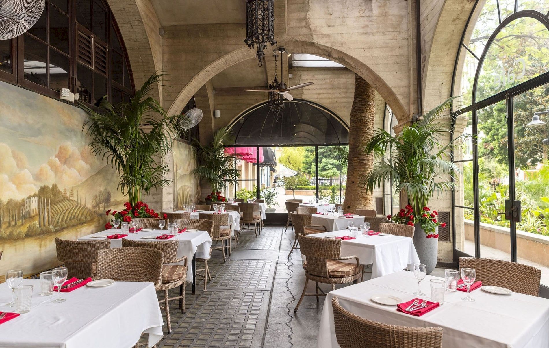 A stylish indoor dining terrace with set tables, palms, arched stone walls, chandelier lighting, and large windows by a mural scene.