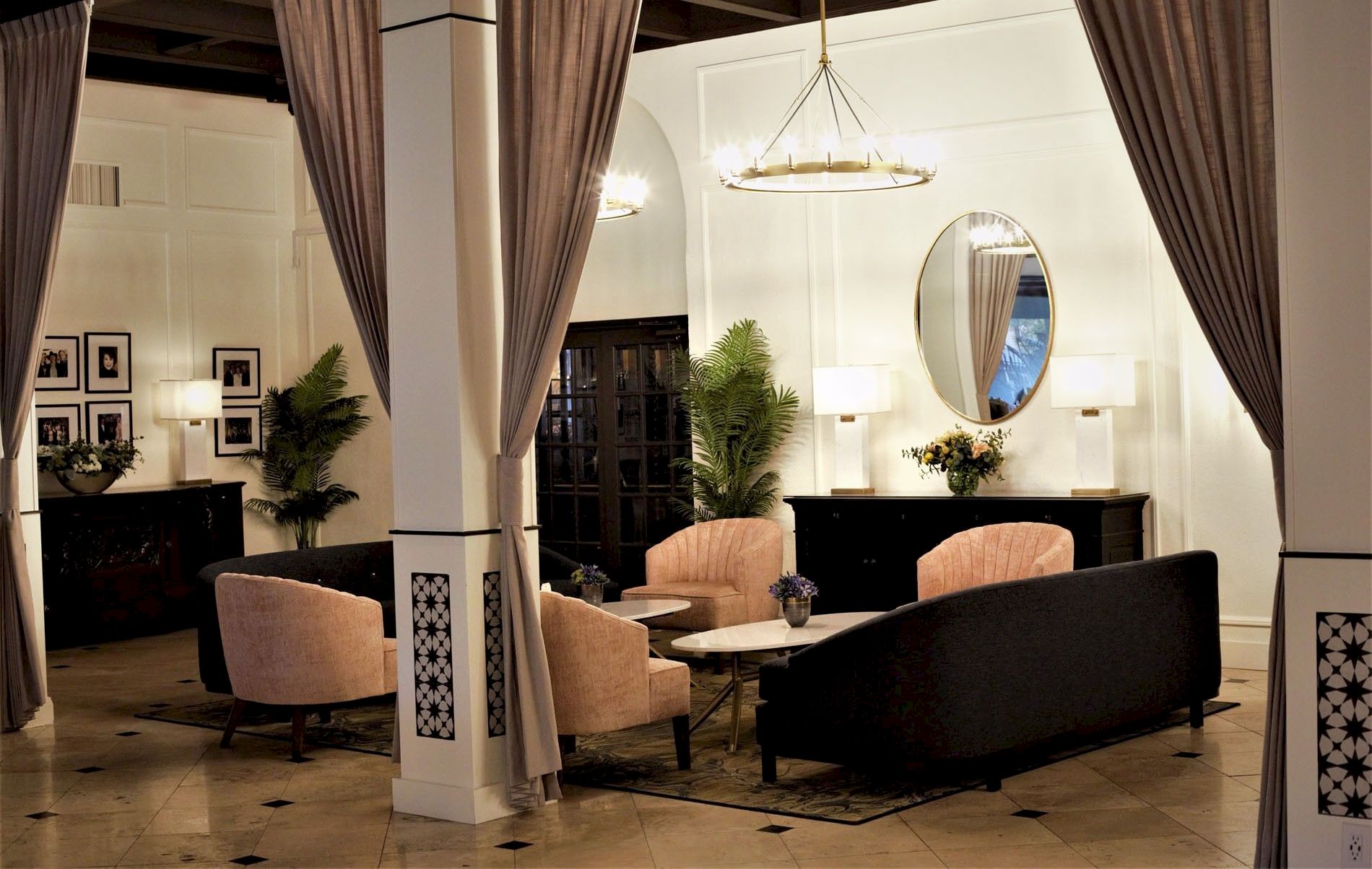 A luxurious living room with tall curtains, plush sofas, a chandelier, a round mirror, and decorative plants, all neatly styled.