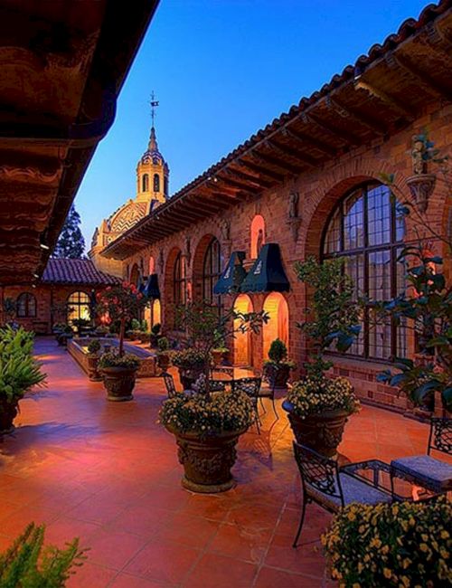 A sunlit Spanish-style courtyard with brick arches, potted plants, and a church bell tower rising in the distance at dusk.