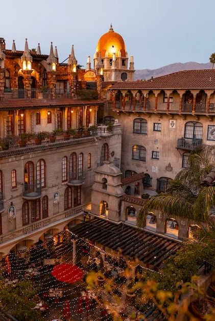 An ornate courtyard at dusk with lanterns and warm lights, surrounded by arched stone buildings and a domed tower.