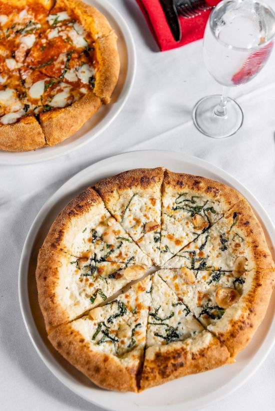 Two pizzas on plates with herbs/cheese, plus a glass of water and red utensils on a table.
