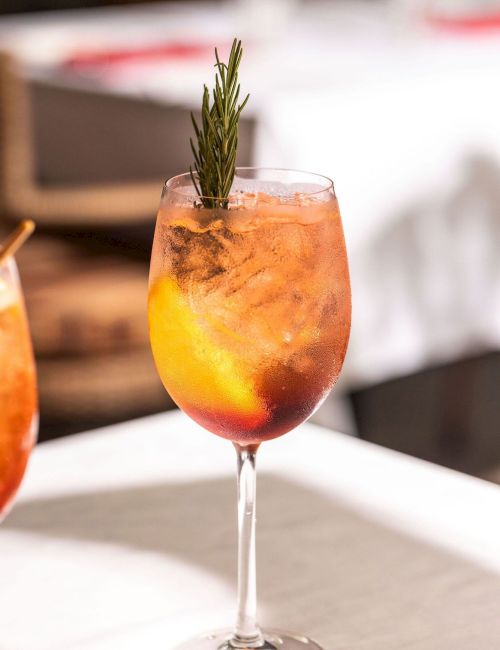 A frosty orange cocktail in a wine glass, garnished with a rosemary sprig and ice, on a patio table.