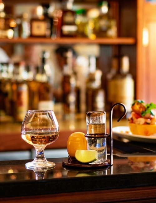 A bar counter scene with a glass of amber drink, a small bottle, and a plated appetizer in a warmly lit pub.