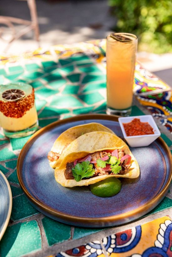 Outdoor table with tacos on a blue plate, chips in a bowl, and iced drinks in tall glasses, on a colorful blanket.