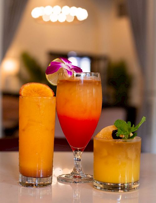 Three colorful cocktails&mdash;orange, red, and yellow&mdash;garnished with fruit and a flower, on a bar counter with a blurred lounge background.