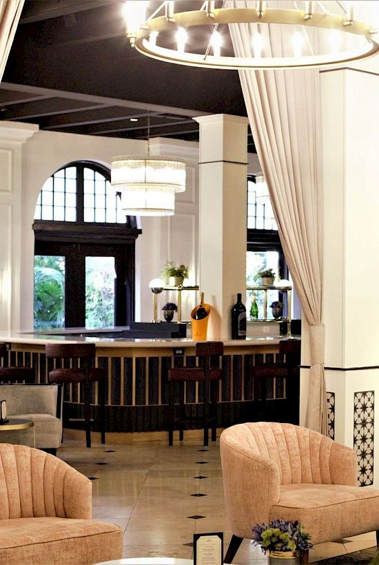 A luxurious hotel lounge with cream curtains, glowing chandeliers, velvet seating, and a central bar under arched windows.