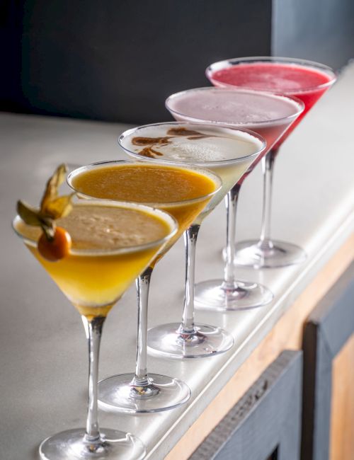 Four layered cocktail martinis in stem glasses&mdash;orange, creamy, clear, and red&mdash;garnished and ready on a tray.