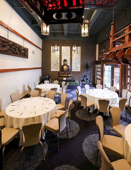 A formal dining room with round tables set for meals, wooden screens, hanging lanterns, and a seated Buddha statue in the center.