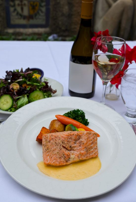 Plated salmon with roasted veggies, salad, and a bottle of wine with a glass topped with red flowers.