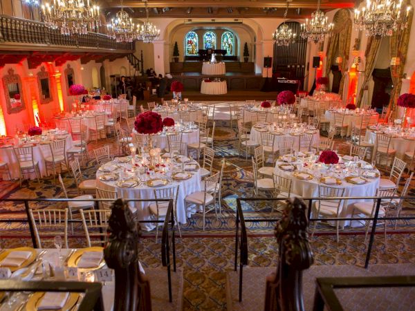 A grand banquet hall set for a formal event, with chandeliers, red lighting, and neatly arranged round tables and chairs.