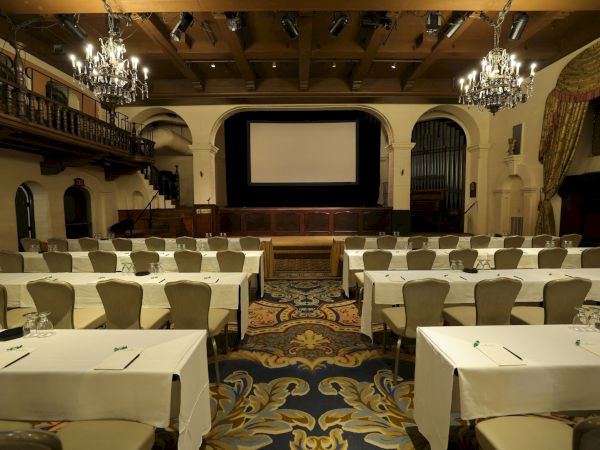 Elegant banquet hall with chandeliers, set tables, arched walls, and an empty stage with curtains.