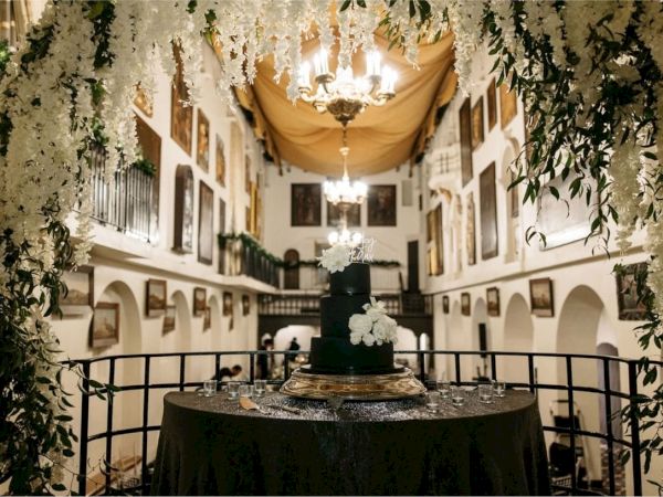 A wedding reception setup: a tiered dark cake on a table, surrounded by white flowers and hanging lights in a grand indoor hall.
