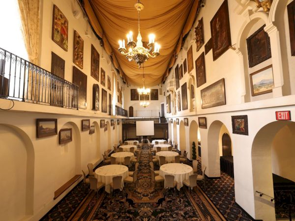 An elegant banquet hall with chandeliers, draped ceiling, framed paintings, round tables, and an EXIT sign along the arches.