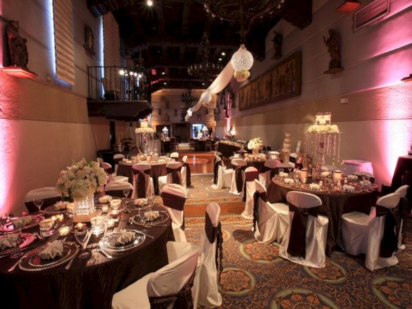 A decorated ballroom wedding reception setup with round tables, white chair covers, chandeliers, candles, pink uplighting, and floral centerpieces.