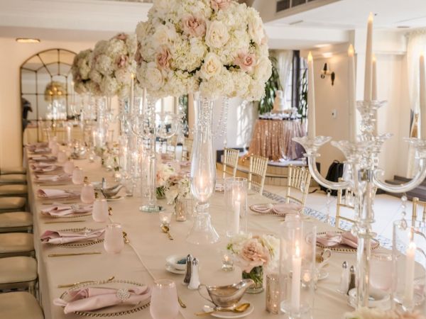 A lavish wedding reception table setup with white-and-cream floral centerpiece, crystal glassware, candles, and gold chairs, glowing warmly.