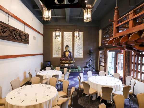 A fine-dining room with round tables set for meals, a large seated Buddha statue at the back, and dramatic red artwork on the ceiling.