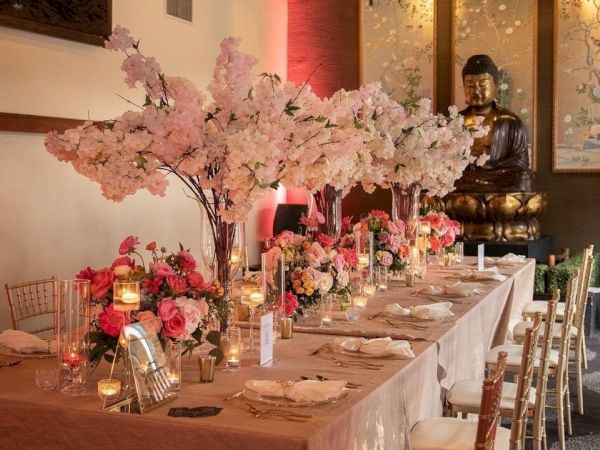 A formal event table set with pink cherry-blossom flowers, candles, and elegant chairs in front of a Buddha statue backdrop.