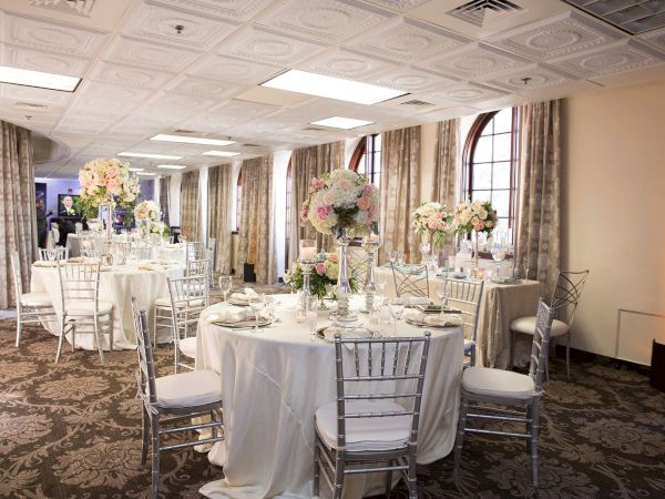 A formal banquet hall with round tables set for a wedding or event, decorated with white cloths, silver chairs, and large pink-and-white floral centerpieces.