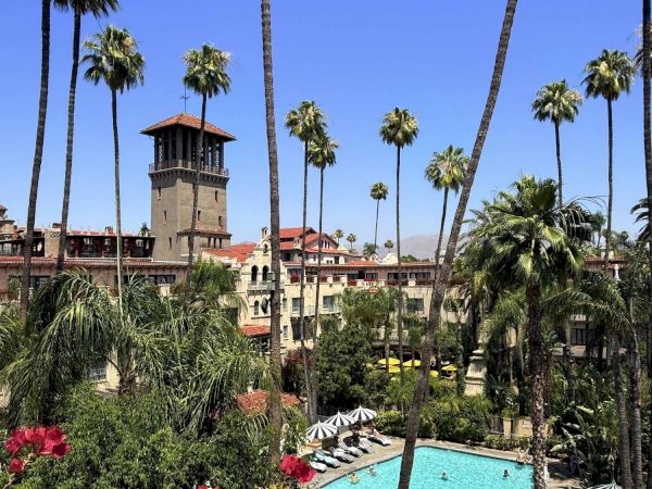 A sunny tropical resort scene with towering palm trees, a bright blue swimming pool, manicured gardens, and red flowers beneath a clear sky.