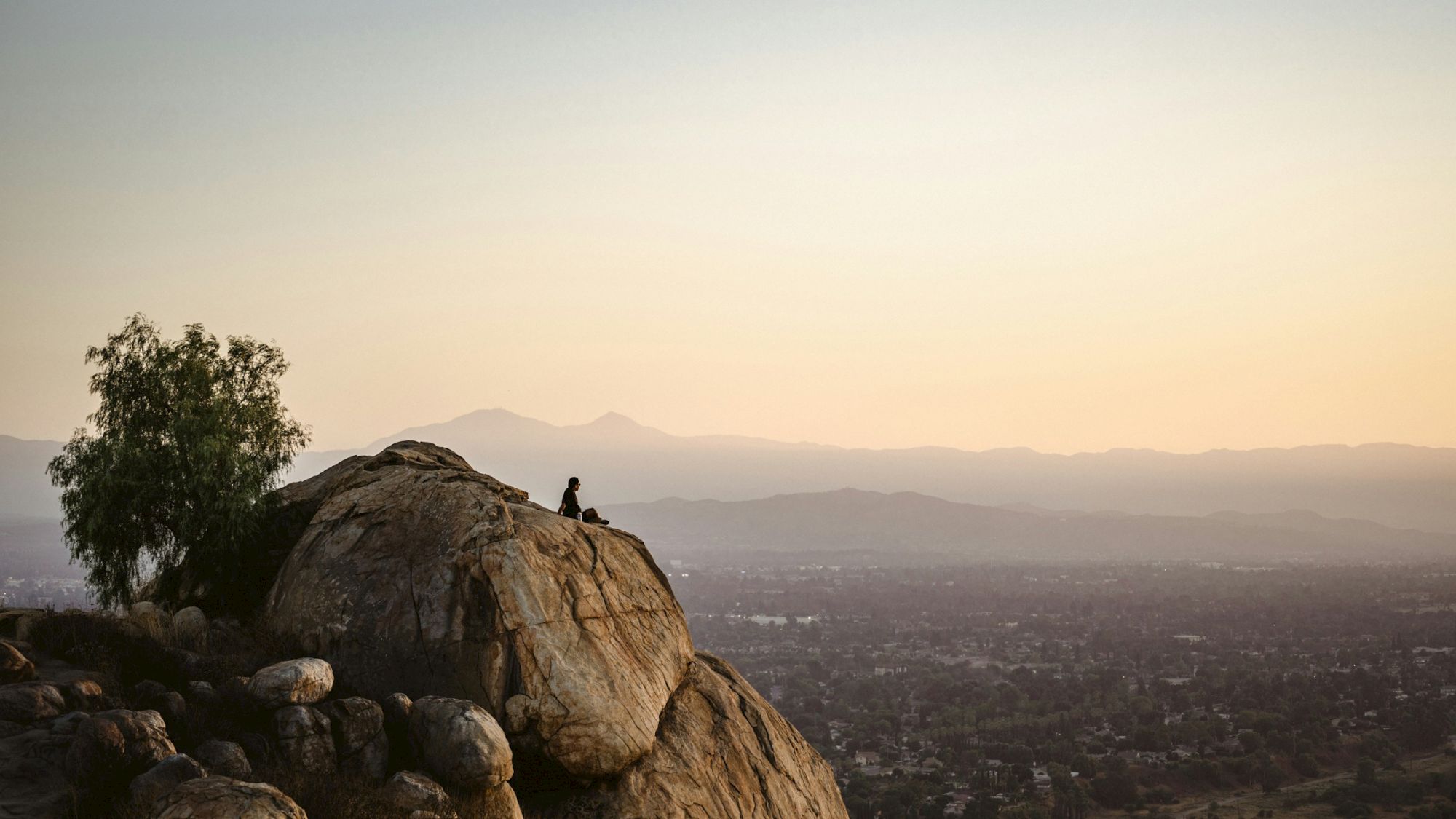 A lone person sits atop a huge boulder, overlooking a distant city and hazy mountains at sunset.