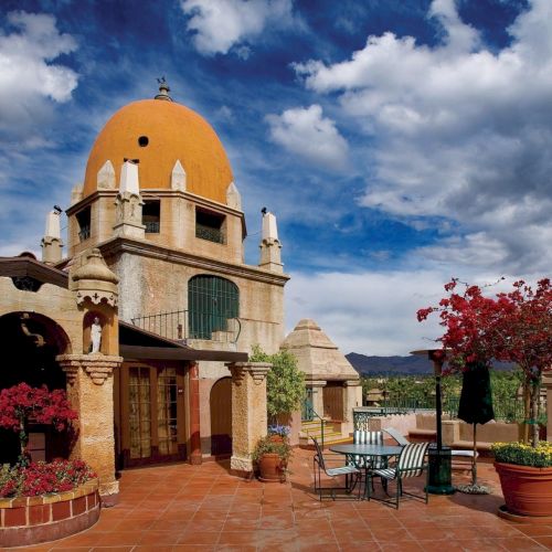 A sunlit courtyard with red flowers and patio chairs, framed by a church-like building with a towering yellow dome under cloudy skies.